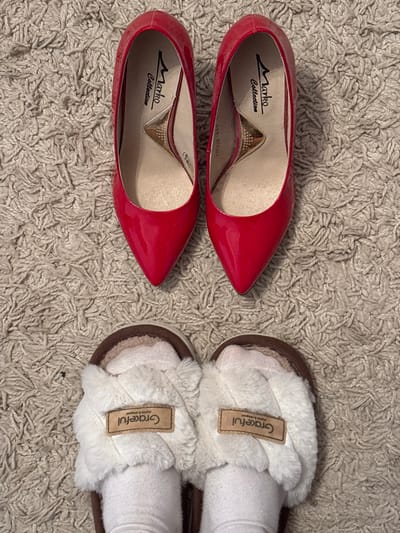 The scene, situated in the city of Velikiye Luki, Russia, depicts footwear arranged on a light-colored, textured carpet. In the upper half of the frame, a pair of red, pointed-toe, patent leather high-heeled pumps are visible. Their insoles display "Marko Collection" branding with a distinctive gold-colored insert. Below these pumps, a person's feet, wearing white socks, are observed inside a pair of brown and white furry slippers. Each slipper is adorned with a tan-colored tag featuring the word "Graceful".