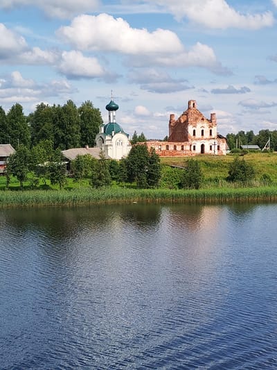 The image depicts a scene in Ankhimovo, Russia, featuring a body of water in the foreground with ripples reflecting the sky and surrounding landscape. On the bank of the water, there is a line of tall green reeds. Behind the reeds, a verdant grassy slope rises, dotted with trees and some buildings.
Dominating the midground are two structures that appear to be churches. To the left, a white church with a green dome and a cross is intact and appears to be in use. To the right, a larger, older building, also likely a church, is in ruins. It is constructed of red brick, with missing sections of walls and roof, and its windows are empty frames. A cross stands in front of the ruined church.
Further back, there are more trees and smaller buildings, including what looks like a house with a grey roof and a simple, modern-looking structure with a pitched roof. The sky above is blue with fluffy white clouds. The overall impression is a pastoral landscape with historical and religious architecture.