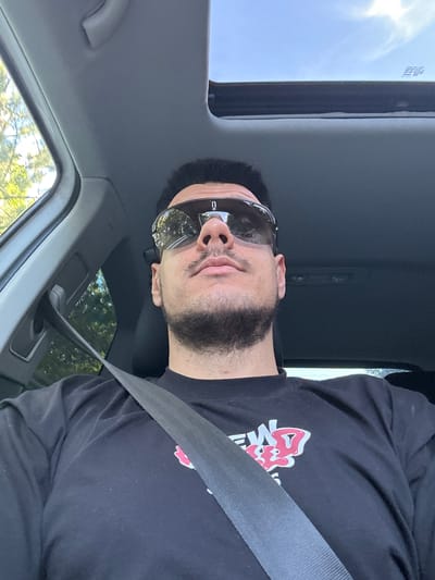 The media file shows a person wearing sunglasses and a black t-shirt with a red and white graphic design, possibly a logo or artwork. The person is inside a car, as evidenced by the seatbelt, car interior details like the sunroof, and the view of trees and foliage through the windows. The image is taken from a low angle, looking up at the person. The lighting suggests it is daytime, with sunlight coming through the sunroof. The person's expression, with closed eyes and slightly parted lips, could indicate relaxation or a moment of repose. The background visible through the windows suggests a rural or suburban setting with green trees. The overall mood is casual and relaxed.