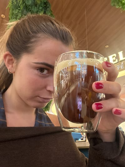 A woman with fair skin and brown hair is holding a glass of dark liquid, likely iced coffee, in front of her face. Her nails are painted a dark red color. She is wearing a plaid shirt and a brown sweater. She is looking at the camera with a neutral expression. In the background, there are green plants and a sign that says "CAFE." The scene is likely inside a café. The café may be located in Senhora da Hora, Portugal, but this is not evident from the image.