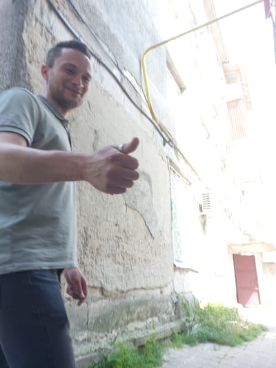 A man, appearing to be in his late twenties or early thirties, stands outdoors in a city. He is wearing a grey polo shirt and dark trousers. He is looking towards the camera and has his right arm extended forward, giving a thumbs-up gesture. He is wearing a silver ring on his left hand. The background consists of a textured building wall on the left and a brightly lit building with a red door on the right. Overhead, a yellow pipe runs diagonally across the scene. The ground appears to be paved with grey tiles or concrete, with some greenery growing at the base of the wall and alongside the paved area. The lighting suggests it is daytime, with strong sunlight creating some overexposure in the right side of the image. The scene is likely in Edineț, Moldova, given the architectural style and the presence of outdoor elements common in Eastern European cities.