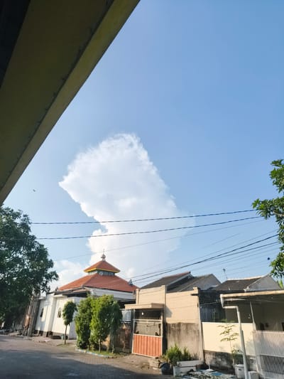 The image shows a street scene in the city of Barengkrajan, East Java, under a clear blue sky with a large, fluffy white cloud formation. On the left side of the street, a white building with an orange-tiled roof and a dome, likely a mosque, is visible. Several green trees are planted in front of the building. Adjacent to the mosque are several residential houses with dark tiled roofs. Some houses have fences and gates. Power lines are strung across the sky, running between poles. The street itself is paved with asphalt. In the upper left corner, a portion of a concrete structure, possibly a bridge or overhang, is visible. A small bird can be seen flying in the sky to the left of the large cloud.