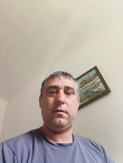 This is a medium shot, waist-up selfie of a man with short, graying hair, dark eyebrows, and light skin. He is looking directly at the camera with a neutral expression. He is wearing a dark gray t-shirt.

In the background, a light-colored wall is visible, with a painting hanging on it. The painting appears to be a landscape, possibly depicting a winter scene with trees and water. The wall has some faint cracks and imperfections. The lighting suggests an indoor setting, likely during the daytime. The overall scene is simple and personal, giving the impression of a casual moment captured in a home environment. There is no visible text in the image.