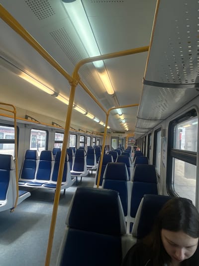 The image depicts the interior of a commuter train carriage, likely in Aprelevka, Russia, with a focus on the empty blue and white seats. The carriage is well-lit by fluorescent lights on the ceiling, and yellow handrails are prominently featured throughout, indicating a public transportation setting. The windows show glimpses of a station platform, with some signage and a person standing outside, suggesting the train is either arriving or departing. In the foreground, a young woman with dark hair is seated, looking down, her face partially obscured. The overall impression is of an ordinary journey, capturing a moment of quiet transit within a functional, modern train carriage.