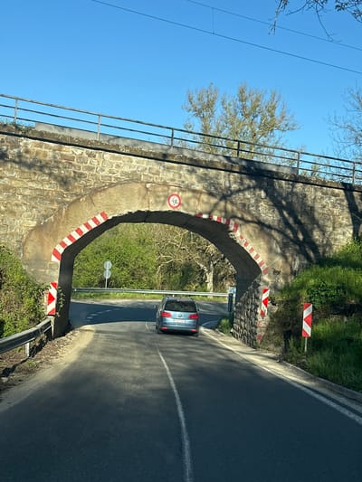 This daytime image depicts a stone arch bridge spanning over an asphalt road in a rural or semi-rural setting near Bilka, Bulgaria. The road curves slightly as it passes beneath the underpass.
A silver passenger car, with a partial license plate "BX" visible (suggesting the Varna region of Bulgaria), is seen driving away from the viewer, having just passed through the archway. The bridge is constructed from irregular stone blocks. Its inner arch is marked with a distinctive red and white diagonal stripe pattern, emphasizing the opening. A circular red and white sign indicating a "4.0" meter height restriction is attached to the archway. Above the bridge, a metal railing runs along its top edge, set against a clear blue sky.
Bright sunlight casts sharp shadows, including intricate patterns of tree branches on the stone surface of the bridge. Green vegetation, including trees (some with bare branches, suggesting late winter or early spring) and grassy areas, surrounds the road and bridge. Several red and white striped chevron signs, serving as hazard markers, are positioned along the roadside approaching and under the bridge. Power lines are visible in the upper part of the frame. The road features a white dashed lane marking. The overall scene suggests a sunny day with good visibility.