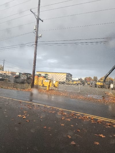 A gloomy overcast day reveals a street with scattered brown leaves. To the side is a chain-link fence protecting a yard filled with heavy equipment, mostly yellow. A tall yellow warehouse-like building is visible in the background. An aged wooden utility pole stands prominently, supporting multiple power lines stretching across the sky. The street surface is wet, reflecting the muted light.