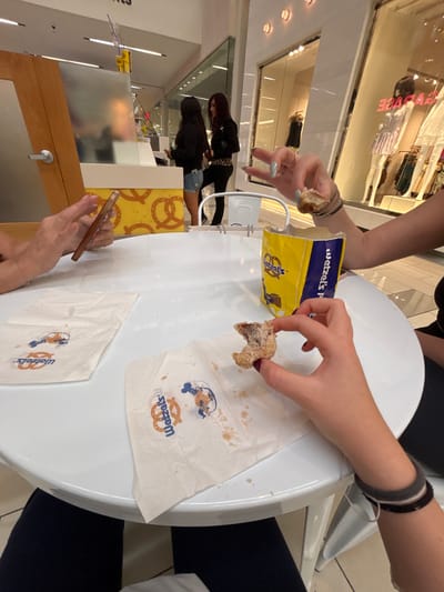 The image depicts an interior scene within a commercial shopping mall in Culver City, United States of America. In the immediate foreground, a white circular table is shown from a seated perspective. On the left side of the table, a person's hands are visible, holding and operating a light-colored smartphone. On the right side, another person's right hand holds a partially consumed, sugar-dusted pretzel bite over a "Wetzel's Pretzels" napkin that displays the brand logo and a Mickey Mouse pretzel graphic, alongside residual sugar and crumbs. A yellow "Wetzel's Pretzels" paper bag, containing additional pretzel bites, sits on the table between the two individuals.
In the mid-ground, a "Wetzel's Pretzels" counter is positioned. Its base features a yellow and brown pattern with pretzel motifs. Two women are standing at this counter. The woman on the left, with dark hair, wears a dark long-sleeved top and light denim shorts. The woman on the right, possessing reddish-brown hair, is dressed in a dark jacket, a leopard-print garment (possibly a top or accessory), and dark pants.
The background reveals a well-lit mall environment with white walls, recessed ceiling lighting, and reflective glass storefronts. One visible storefront clearly displays the word "GARAGE" and features mannequins dressed in various retail clothing items. A wooden door with a silver handle is partially visible to the left of the pretzel stand.