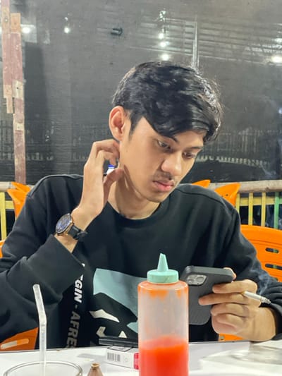 A young adult male is depicted seated at a white table, presumably in a casual dining establishment located in Alue Mudem, Indonesia, under low-light conditions consistent with nighttime. He wears a black long-sleeved shirt with a light blue graphic design partially visible on the chest and a black-strapped wristwatch with a rose gold-colored bezel on his left wrist. His right hand is raised, with fingers making contact with his neck/jawline. His left hand holds a black smartphone with a textured case, and his gaze is directed downwards towards the phone's screen. A lit cigarette, emitting smoke, is held between the fingers of his left hand. On the table directly in front of him are a clear plastic squeeze bottle with a light blue cap, containing a red liquid (likely sauce), and a partially visible white pack of cigarettes. A clear glass containing a white straw is visible on the far left of the table. The background is blurred and dark, revealing orange chairs and light-colored wooden lattice structures, with reflections from overhead lights.