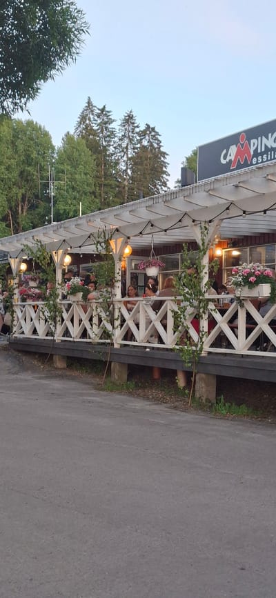The image shows an outdoor seating area of a restaurant or cafe, identified by the sign "CAMPING Messilä" at the top right, indicating the location is Salpakangas, Finland. The area is covered by a white pergola decorated with hanging lights and climbing plants. White wooden railings with a criss-cross pattern line the elevated deck where patrons are seated at tables. Several people are visible, some eating and drinking, others engaged in conversation. Potted flowers, primarily pink and white, are placed along the railing and on tables, adding color to the scene. The background consists of green trees and a clear sky, suggesting a daytime or early evening setting. A paved area, likely a road or walkway, is in the foreground.