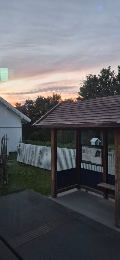 This media file shows an outdoor scene at dusk, likely in Biri, Norway, given the context. The sky is filled with wispy clouds, painted with soft hues of pink, orange, and gray, indicating the end of daylight. A wooden roofed structure, possibly a bus stop or shelter, is a prominent object in the foreground, supported by thick wooden posts. Inside the shelter, a dark blue panel is visible, with a white poster or sign featuring text and a QR code. To the left of the shelter, a white building with a sloped roof is partially visible, and beyond that, trees with dense foliage add to the natural setting. A white picket fence runs across the middle ground, separating the area around the shelter from a grassy patch dotted with small flowers. A darker, asphalt-like surface occupies the right side of the frame, suggesting a road or pathway. There are no people visible in the frame, and the overall mood is tranquil, with the soft light of dusk creating a peaceful atmosphere. The visible text on the poster appears to be in English, with "EN" prominently displayed.