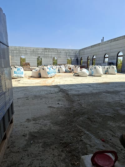 The image depicts an outdoor construction site in Kiryat Gat, Israel, under a clear blue sky. The foreground shows a rough, concrete ground surface with scattered debris. To the left, a pallet of building materials is partially visible, wrapped in plastic. In the middle ground, several large, white bags, likely containing construction materials, are arranged on the ground. Some of these bags have blue lettering, including Hebrew text and "02-991-0771". Behind the bags, a low, unfinished concrete wall forms a corner of a building, with arched openings revealing trees and foliage beyond. A metal ladder leans against the wall near the openings. In the background, the wall continues, with two dark pipes protruding from its top edge. The scene suggests ongoing construction work.