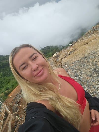 A blonde woman with light skin and a red bikini top and black jacket takes a selfie. She is smiling and looking at the camera. The background is a steep, rocky hillside with green vegetation in the distance, partially obscured by fog. The location appears to be a construction or excavation site. The weather is overcast. The photo was taken outdoors.