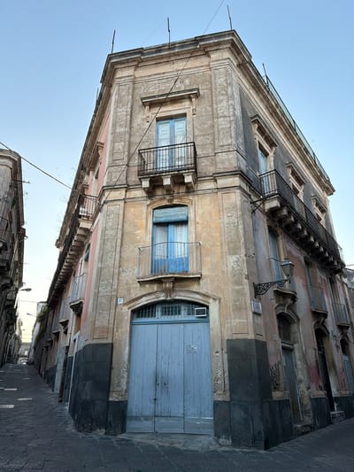 The photograph captures a street scene in Acireale, Italy, focusing on the architecture of a building along a narrow cobblestone street. The building's facade exhibits a mix of aged and weathered surfaces, with a color palette dominated by gray, beige, and hints of pastel blue on window shutters and door panels.

Architectural details include ornate window frames, small wrought-iron balconies, and a fire escape ladder clinging to the side of the building. Wires crisscross the sky above. At street level, a large blue door anchors the base of the structure. A street number '27' is visible on the facade beside the door. The perspective is from a low angle, accentuating the height of the building and its position within the tight streetscape. The sky is light blue.