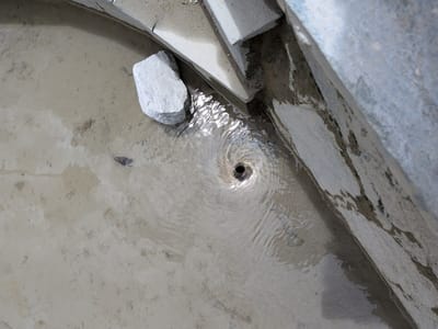 The image shows a drain where water is swirling into a funnel shape. The drain is located in a sandy area with some concrete structures around it. There is a rock to the left of the drain. The water is murky and appears to be dirty. There are no people or animals visible in the image. The location is identified as Panamik, India.