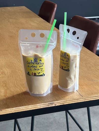 The image displays two clear plastic stand-up pouches, each containing a light brown, milky beverage and a bright green plastic straw. Both pouches feature a yellow rectangular label with black text, prominently reading "CAFFEINATED HUMAN OF EL PASO," a QR code, and various small illustrations including people and a coffee cup logo. One label includes "@1031Cafe." The pouches are positioned on a light-colored wooden table with visible wood grain and dark metal legs. In the blurred background, dark-colored chairs with reddish-brown upholstered backs are discernible against a dark wall. The scene is an interior setting, likely a cafe or commercial establishment in El Paso, United States, as indicated by the labels. No individuals are present, and all objects are stationary.