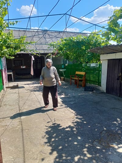In Salhany, Ukraine, on a sunny day with scattered clouds, an elderly woman stands on a concrete courtyard. She is wearing a light-colored, ribbed sweater and dark trousers, with brown shoes. Her hair is short and gray, and she appears to be smiling faintly as she looks towards the camera.

The courtyard is bordered by buildings and a green wooden fence. To the left, a building with a corrugated roof has a window and a door, with a sink visible outside. A table and chair are also present. To the right, a wooden bench sits against the green fence. Beyond the bench, a dark wooden door with a lock is visible on another building.

Above the courtyard, a metal structure supports grapevines, casting dappled shadows on the ground. The shadows from the woman and the grapevines create a dynamic pattern on the concrete. The time of day is likely midday, given the bright sunlight and the position of the shadows. The overall scene suggests a rustic, rural setting. There is no visible text in the image.