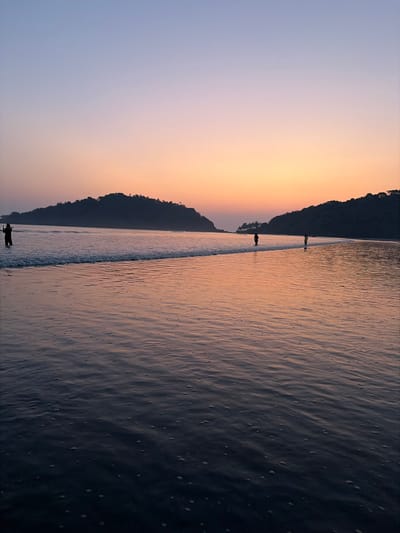 The image portrays a sunset scene at a beach in Kolamb, India. The sky graduates from a light lavender-blue at the zenith to warm orange and pink hues near the horizon. The ocean foreground is dark, reflecting the sky's colors, with subtle ripples and scattered white foam evident on the surface. A distinct breaking wave extends across the mid-ground. In the background, two forested landmasses are silhouetted against the sunset: a larger, rounded island to the left, and a smaller landmass to the right, forming an inlet or bay.

Three human figures are present in the water. On the far left, one individual stands partially submerged, approximately knee-deep, holding an object, likely a mobile device, raised as if capturing an image of the sunset. A second individual, silhouetted and facing away from the viewer, walks in the water further offshore towards the right side of the image. A third, smaller silhouetted individual walks closer to the right shoreline, also in the water. All individuals are partially submerged in the shallow ocean water. No direct interactions between the individuals are discernible.