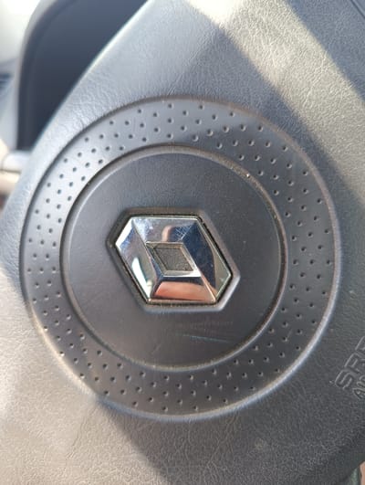 The image displays a close-up view of a dark grey, textured car steering wheel's airbag cover. Centrally located on the cover is a silver-colored Renault logo, a stylized diamond or rhombus shape, which exhibits multiple surface scratches and reflects ambient light and a partial human form in its lower section. Surrounding the emblem is a slightly raised circular ridge, which transitions into a wider circular band featuring a pattern of uniformly spaced small, circular perforations. The texture of the steering wheel material appears to be a grained synthetic leather or plastic. In the lower right quadrant, the partial letters "SRS AIRBAG" are visible, embossed into the material. Dust particles are discernable on various surfaces, particularly within the perforations and around the emblem. Strong, directional light, likely sunlight, illuminates the scene from the upper left, creating distinct highlights and shadows across the textured surfaces. The image provides no visual information to corroborate the specified location in the city of Maiaky, Ukraine.