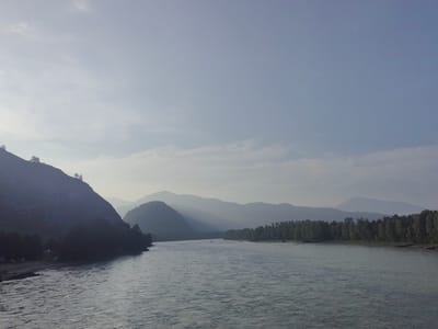This image depicts a wide, calm river flowing through a mountainous landscape in Anos, Russia. The sky is overcast with a light haze, casting a soft glow over the scene. On the left side of the river, a steep, dark mountainside rises, sparsely covered with trees. A small beach is visible at the base of this mountain. On the right bank, a dense forest of green trees stretches along the river's edge, with rolling, hazy mountains visible in the distance. The water's surface is rippled, reflecting the muted light of the sky. No people or vehicles are present in the immediate foreground or along the visible riverbanks. The overall impression is one of natural tranquility and serene beauty.