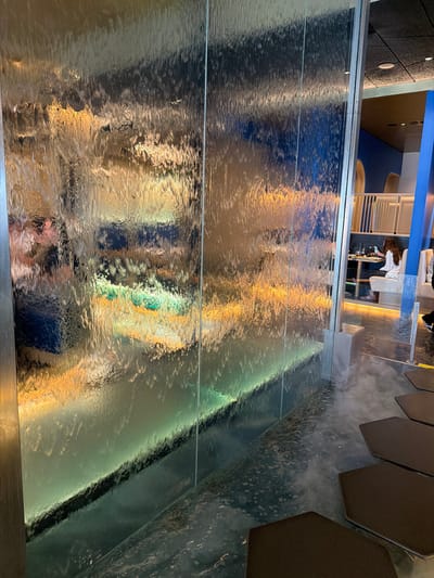 The image shows a water feature or wall in a public area, likely indoors. The water cascades down a multi-paneled surface, creating distorted reflections of the surroundings. Reflected in the water are elements of a dining area, including chairs and tables, and possibly people. The environment suggests a modern or contemporary design, possibly a restaurant or hotel in Torreforta, Spain.