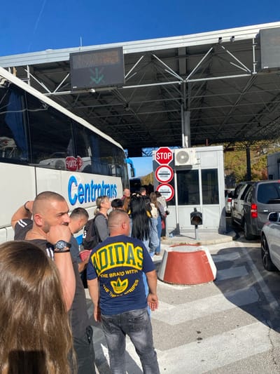 The image depicts a busy daytime scene at a border crossing in Grab, Bosnia and Herzegovina. A white Centotrans passenger bus occupies the left lane, its side reflecting a red "STOP" sign. To its right, a queue of approximately 15-20 individuals, dressed in casual attire including a man in a dark blue Adidas t-shirt, stands facing a grey checkpoint booth. This booth displays a red octagonal "STOP" sign, a circular "CARINA DUANE" (Customs) sign, and a circular "POLICIJA POLICE" sign. An air conditioning unit is mounted on the booth's right side.

Above the lane, a large, metallic truss canopy structure covers the area, supporting an electronic LED display that shows a green downward arrow. In the foreground, a red and white concrete barrier sits on asphalt pavement marked with white lines. To the right of the checkpoint booth, a separate lane features a line of cars, including a dark grey SUV or minivan and other vehicles, waiting to proceed. Hills with sparse vegetation are visible in the background under a clear blue sky.