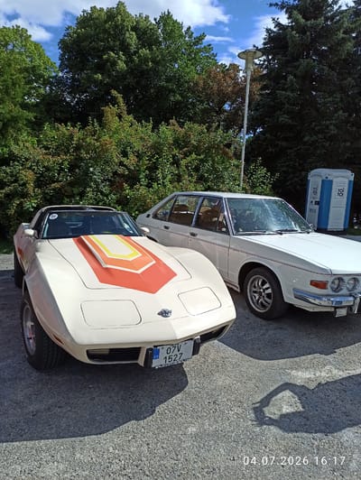 Two vintage cars, a cream-colored Chevrolet Corvette and a white Tatra, are parked on asphalt. The Corvette, in the foreground, has a distinctive orange, yellow, and red stripe pattern on its hood. The Tatra, to the right, is a sedan with a red stripe along its side. In the background, dense green trees and a blue and white portable toilet are visible. The sky is blue with scattered clouds. A lamppost stands to the left of the Tatra. The image is dated 04.07.2026 16:17. The scene appears to be in Mariánské Lázně, Czechia, likely at an outdoor event or gathering of classic cars.