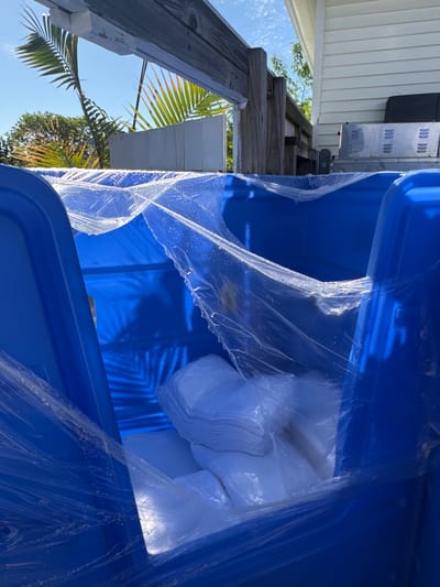 The image shows the interior of a large, blue, plastic container, likely a storage tote or bin. The container is filled with a large bundle of white, plastic-wrapped items, appearing to be stacks of folded white sheets or linens. The container is partially covered with clear plastic wrapping, possibly to protect the contents during storage or transport.

Outside the container, the scene includes elements of an outdoor environment. A wooden structure, possibly a porch or deck, is visible, along with palm fronds and trees. A section of a building with white siding is also present.

Given the presence of palm trees and the building's architectural style, it is plausible that the image was taken in a coastal area or tropical climate. Without additional information, pinpointing the exact location within Edgartown, United States of America, is not possible.