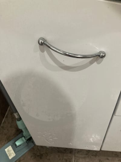 The image shows a close-up of a white cabinet with a silver curved handle. The cabinet appears to be in a domestic setting, possibly a kitchen or bathroom, given the presence of a mop on the floor in the foreground. The floor is tiled with a brown, patterned tile. There is a faint circular mark on the cabinet, which may be a water stain or a shadow. The lighting suggests an indoor environment. The location provided, Козенскі сельскі Савет, Belarus, is not visually represented in the image.