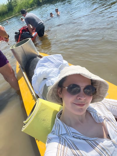 In a shallow, muddy body of water, likely a river or lake, several individuals are engaged in recreational activities on a sunny day. A woman wearing a sun hat and sunglasses is positioned in the foreground, taking a selfie from a bright yellow kayak. She is dressed in a striped open-collared shirt. In the middle ground, a person in a gray t-shirt and black shorts is standing in the water, seemingly adjusting equipment on an orange inflatable raft. Two other individuals, appearing to be children, are visible in the water further in the background, along with another person swimming. The environment is characterized by calm water, a visible shoreline with green vegetation, and a clear sky. The general impression is one of a relaxed, outdoor gathering.
