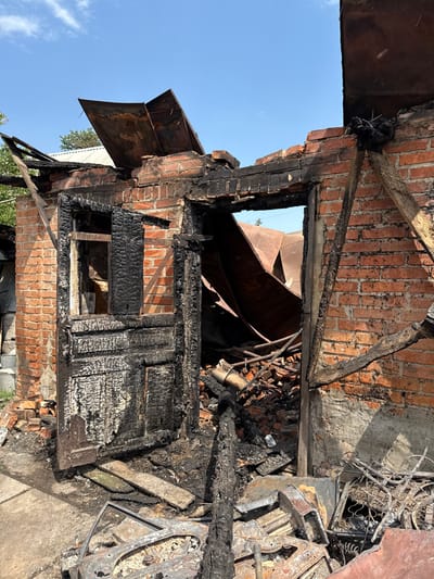 The image displays a severely fire-damaged brick structure in Nedryhailiv, Ukraine. The building's red brick walls remain partially standing but show extensive charring and soot discoloration, particularly along the top sections and around openings. A heavily charred wooden door, fractured and blackened, is partially ajar in the main entranceway. A window opening to its left is devoid of glass, with its wooden frame entirely burnt away. The roof has collapsed, with large, warped, and discolored corrugated metal sheets resting haphazardly within the structure's interior. The ground inside the building is covered with rubble consisting of shattered bricks, fragmented charred wood, and twisted metal debris. Outside the structure, on a concrete or paved surface, additional charred materials are present, including a skeletal metal frame resembling a car door or vehicle component. Charred wooden beams and supports are visible within and around the structural openings. The sky above is clear blue with minimal cloud cover. No individuals or living beings are present in the scene.
