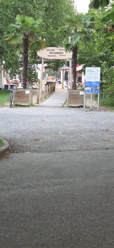 The image depicts a pathway leading into a park or campground area, with lush green trees and palm trees framing the scene. A large, oval wooden sign with multilingual greetings hangs from a tree, reading "WELCOME WILLKOMMEN BIENVENU BENIVENUTO BENVENUTO BIENVENIDO". To the right of the path, a paved walkway continues, bordered by a gravel area. In the background, several people are visible relaxing on lounge chairs under a canopy, with lounge chairs and other recreational items scattered around. A notice board with text and images stands near the entrance, further suggesting a public or recreational space. A bicycle is parked on the grass to the left, near some stored items. The scene appears to be set during the daytime, with soft lighting suggesting an overcast or diffused sunlight. The overall atmosphere is relaxed and welcoming, characteristic of a leisure or holiday destination. The location is identified as Bregenz, Austria.