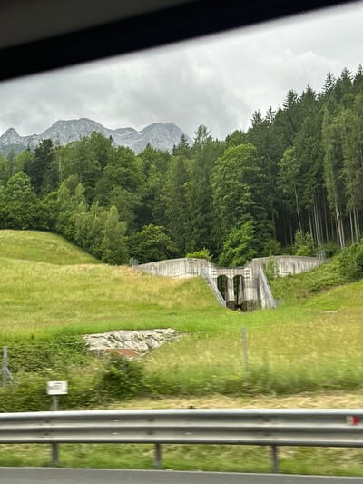 The image depicts a rural landscape in Kuchl, Austria, featuring rolling green hills, a dense forest, and a concrete structure that appears to be an old dam or water management facility. The sky is overcast with clouds. In the foreground, a metal guardrail and a road are visible, suggesting the photograph was taken from a moving vehicle. The concrete structure has two archways and shows signs of wear and tear, partially integrated into the hillside and surrounded by vegetation. The forest behind the structure is thick with evergreen trees. In the distance, hazy mountains can be seen under the cloudy sky. There are no people or animals present in the scene.