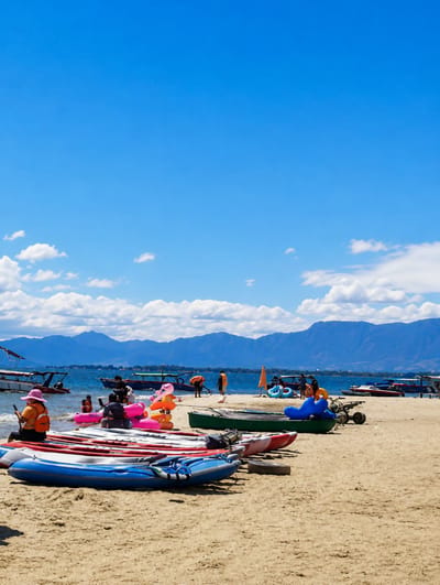 This is a photograph taken at Lumban Bul Bul, Indonesia, a location known for its beach and water activities. The image depicts a sandy beach with several inflatable kayaks and canoes lined up. In the background, the clear blue water of a lake or sea is visible, dotted with various boats. Beyond the water, a mountain range under a bright blue sky with scattered white clouds forms the horizon.
On the beach, there are several people engaged in leisure activities. A woman wearing a pink hat and an orange vest is seated on the sand, looking at her phone. Other individuals, some wearing orange life vests, are scattered around, with some interacting with inflatable toys in the shallow water. A few people are standing near the water's edge, and others are seated on the sand.
The overall atmosphere is one of a sunny day at a recreational beach, with various watercraft available for rent or use. The presence of mountains in the distance suggests a scenic natural environment.