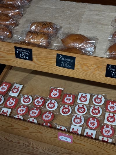 The image depicts a bakery display shelf in Tavrichanka, Russia, showcasing various baked goods. In the upper portion of the frame, loaves of bread are visible, some sprinkled with sesame seeds and packaged in plastic wrap. Below these, a wooden shelf holds smaller items. On the left, a black sign reads "сосиска в тесте 150р" (sausage in dough, 150 rubles). To its right, another black sign says "Капуста 100р" (cabbage, 100 rubles). Further right, a third black sign is partially visible with "Лук 10" (onion, 10) potentially indicating onion-filled pastries. The bottom shelf is filled with individually packaged cookies decorated with a stylized red and white sheep or llama face. A pink price tag at the bottom left corner reads "ПРЯНИК 250р" (gingerbread, 250 rubles). The overall environment suggests a retail setting for baked goods.