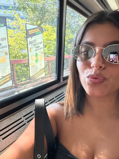 A female subject is captured in a close-up selfie inside a vehicle, likely a bus or train, in New Brunswick, New Jersey. She has dark hair and is wearing oval, silver-framed sunglasses with tinted lenses; a reflection of a mobile phone held for the selfie is visible in the right lens. Her lips are pursed in a "kiss face" expression. She wears a dark, possibly strapless top, with a black bag strap visible on her left shoulder. Through the vehicle's window, an outdoor environment under bright sunlight is visible. Two large, vertical advertisement banners stand prominently. The banner on the right features "Dial 811," "1-800-272-1000," and "www.nj-call.org," displaying a logo depicting the state of New Jersey. The adjacent banner to the left mentions "Law" and a similar contact number. Behind these banners, dense green foliage and a red metal railing are visible, with a clear sky or distant building appearing above. Horizontal ventilation grates are present on the vehicle's interior wall below the window.