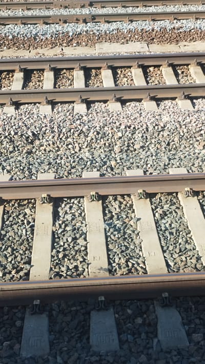 The image shows a close-up, overhead view of multiple sets of train tracks in Vyshny Volochyok, Russia. The tracks are constructed with dark metal rails laid on top of concrete sleepers, which are themselves embedded in a bed of grey and brown gravel ballast. Some of the concrete sleepers have visible markings or inscriptions. The scene appears to be outdoors, with bright sunlight casting shadows on the tracks and ballast. There are no people or vehicles visible in the image. The composition is focused solely on the railway infrastructure.