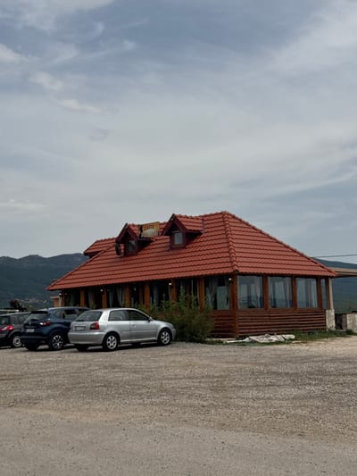 The image depicts a cafe with a red tiled roof and wooden walls situated in the city of Vidrovan, Montenegro. Several cars are parked in front of the cafe on a gravel lot. The cafe has large windows, and a sign that reads "CAFFE POGLEDINE" is visible on the roof. In the background, there are hills and mountains, and the sky is overcast with clouds.