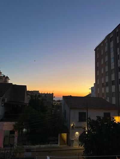 The image depicts a cityscape during a sunset, likely in Agropoli, Italy, given the context. The sky transitions from blue to orange as it approaches the horizon, where a narrow band of water is visible. Buildings of varying heights and architectural styles occupy the foreground. A tall, modern building with multiple stories is visible on the left, while older, shorter buildings with tiled roofs and traditional facades are present on the right. Trees are scattered throughout the scene, obscuring some of the structures. A building with the name "Vecchia" is visible, suggesting a restaurant or establishment with an Italian theme. There are no people visible in the image.