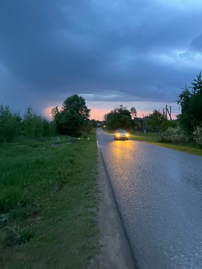 The image captures a rural or semi-rural road scene during twilight or dusk, likely after rainfall. A light-colored car is approaching from the middle distance, its bright yellow headlights illuminating the wet road surface which reflects the light, creating a sparkling effect.
The road is flanked on both sides by abundant green vegetation, including lush grass in the foreground and dense trees and bushes stretching into the background. The sky, which occupies a significant portion of the image, is a deep, cloudy blue-gray, indicating either heavy cloud cover or the onset of night. A narrow band of lighter sky with soft pink and orange hues is visible towards the horizon on the left, suggesting the lingering glow of sunset.
Faint structures, possibly power lines or utility poles, can be glimpsed through the trees on the left side of the road in the mid-ground. The overall impression is one of quiet transit in a natural, somewhat dramatic setting, with the car's headlights cutting through the low light. The wet conditions add to the atmospheric quality. The location context provided is Pyatnitsa, Russia. No visible text is present in the image.