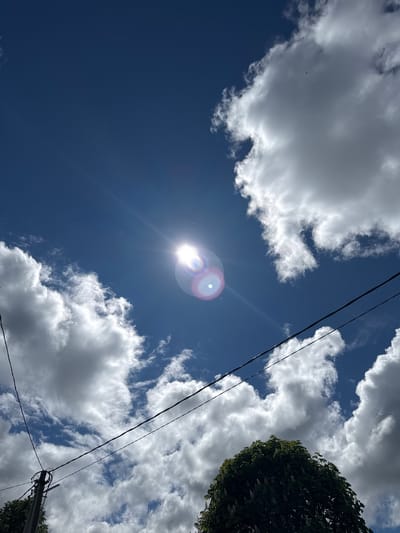 The image is taken from a low angle, looking up into a sky with scattered white clouds against a deep blue backdrop. The sun is visible, appearing as a bright white disc with lens flare effects. Electrical power lines diagonally cross the frame. Part of a tree with green foliage is visible in the top right corner. The specific location in the city of Slobodka, Russia cannot be identified solely from the information in the image.