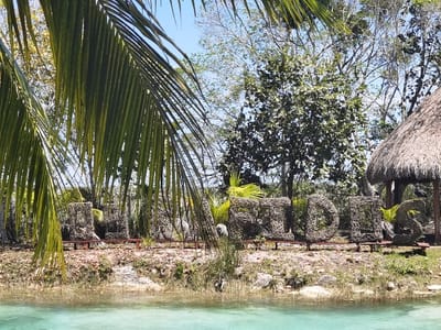 The image shows a "Riders" sign made of twigs and branches, located in front of a body of turquoise-colored water. There is a thatched roof hut in the background, and a palm tree frond obscures much of the view. The scene is taken from the ground level, looking towards the sign. It appears to be a recreational area located in the town of Unidad de Riego, Mexico, near the coordinates latitude: 18.591, longitude: -88.434.