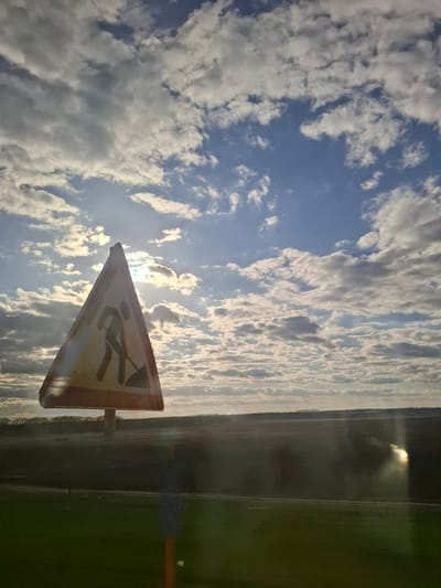 The image displays a landscape scene under a partly cloudy sky, featuring a road work sign in the foreground. The sky occupies the upper portion of the frame, showing vast expanses of blue interspersed with numerous white and grey clouds. The sun is visible, partially obscured by clouds, radiating bright light and creating a noticeable lens flare.

In the lower foreground, a yellow triangular road sign with a reddish-brown edge is prominently displayed. The sign features a black silhouette of a person holding a shovel, commonly signifying "road work ahead" or "men at work." The sign itself appears somewhat weathered. Behind the sign, the horizon stretches across the middle of the image, showing a long, dark strip of land that could be a distant road, forest, or field. The lower-right portion of the image reveals a green, open field or grassy area.

The scene captures a bright, daytime moment, characterized by a mix of sunlight and cloud cover. The presence of the road sign suggests ongoing or upcoming construction or maintenance in the vicinity. The setting appears to be a vast, open, and potentially rural or semi-rural area in Knyazevo, Russia. No specific activity beyond the implied road work is taking place within the visible frame. There is no explicit text visible on the sign, only the symbolic pictogram.