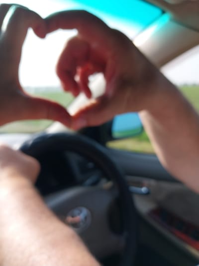 The image shows a close-up, blurred view of two hands forming a heart shape, with fingers touching to create the outline. The hands are the primary subjects, with a suggestion of a person's upper body and arms. The setting appears to be inside a vehicle, indicated by the presence of a steering wheel and parts of the car's interior, such as the dashboard and window. The lighting suggests it is daytime, possibly sunny, due to the bright white background and illuminated areas. The focus is on the gesture of the hands, implying an action of love, affection, or connection. There are no visible text elements in the image. The overall impression is intimate and personal, captured within the context of a car journey.