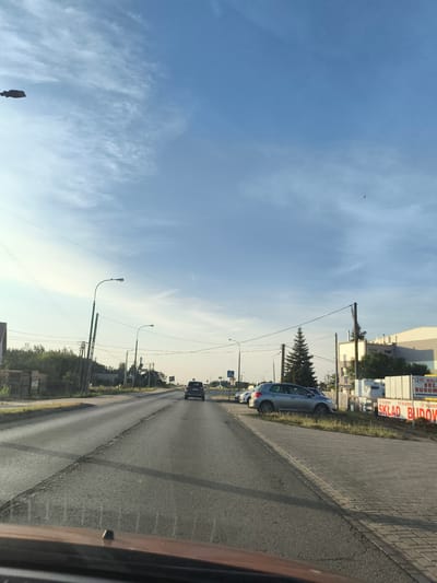 The image captures a daytime scene on a two-lane asphalt road in Czosnów, Poland. The road appears to be an arterial route with parked cars and buildings lining the right side. The sky is a clear blue with scattered wispy clouds.
On the left side of the road, there are stacks of what appear to be building materials, possibly lumber or palettes. Further down the road, there are trees and some low-rise buildings. Several utility poles with electrical wires are visible along the roadside.
On the right side of the road, there is a sidewalk paved with interlocking bricks. Parked cars are visible along this side, including a silver hatchback and a dark-colored SUV. A large, white building with some signage is also present. One sign, in red lettering, reads "SKŁAD BUDOWY" which translates to "BUILDING SUPPLY STORE," and "MALINOWSKI". There is a tall, dark green evergreen tree in front of this building.
The perspective suggests the image was taken from inside a vehicle, looking forward. The road surface shows some wear and tear, with cracks visible in the asphalt. The overall impression is a commercial or semi-industrial area on the outskirts of a town.