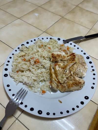 The image displays a close-up view of a meal served on a white circular plate with a black-dotted rim. The plate holds a portion of white rice mixed with diced orange vegetables, consistent with carrots, and minor green specks, occupying the left side. To the right, several pieces of cooked chicken breast are arranged, exhibiting light browning and char marks from grilling or searing. A silver-colored dinner fork with a black handle is positioned in the lower-left, partially resting on the plate's rim and the underlying surface. A matching silver-colored dinner knife with a black handle is visible in the upper-right, also partially on the plate's rim and the surface. The surface beneath the plate and cutlery consists of light tan or beige square ceramic tiles, laid in a diagonal pattern relative to the camera, with darker grout lines. A faint white mark is present on one of the upper tiles. No individuals are depicted, and no explicit actions are occurring. The specific location within the city of Covilhã, Portugal, cannot be determined from the visual information presented in the image.