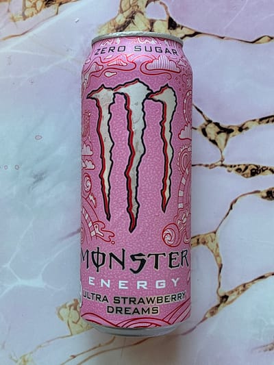 A pink Monster Energy can is photographed from a high angle, centered within the frame. The can features the Monster logo in white, outlined in black and red, with three claw marks. Above the logo, "ZERO SUGAR" is printed in white text with red accents. Below the logo, "MONSTER ENERGY" is written in black text, followed by "ULTRA STRAWBERRY DREAMS" in white text with red accents. The can has water droplets on its surface. The background is a marble-like pattern in pink and white with brown veining. The scene is indoors. The location is not identifiable within the city of Haapsalu, Estonia.