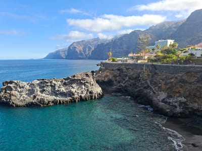 This is a landscape photograph taken from a high vantage point overlooking the coast of Santiago del Teide, Spain. The scene is dominated by dramatic cliffs that rise steeply from the ocean. The water is a vibrant turquoise near the shore, transitioning to a deep blue further out. A large, jagged rock formation juts out of the sea in the foreground.
On the cliffside, a resort hotel with white walls and blue-tinted windows is visible, along with other white buildings with red-tiled roofs. These structures are surrounded by lush green vegetation, including palm trees. A stone wall runs along the cliff edge, providing a boundary and pathway. Steps carved into the rock face lead down towards the water. The sky is a clear blue with scattered white clouds, and sunlight illuminates the scene, casting shadows on the cliffs. The overall impression is one of a scenic coastal resort area with rugged natural beauty.