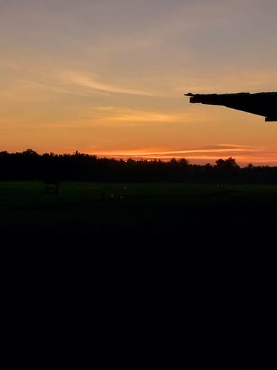 The image shows a silhouetted view of a rural landscape in Labissa, Indonesia, during sunset. The sky is a gradient of orange and purple, with thin, wispy clouds. In the foreground, a dark silhouette of a roof edge extends from the right side of the frame. Below the sky, a line of dark trees forms the horizon. In front of the trees is a field of dark green crops. A small, dark structure, possibly a shelter or hut, is visible in the middle ground of the field, along with several faint, indistinct human figures. The bottom portion of the image is completely black, obscuring any details in the immediate foreground.