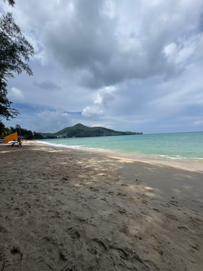The image shows a wide, sandy beach with gentle waves lapping the shore under a cloudy sky. In the foreground, the sand is textured with footprints and debris, cast in dappled sunlight. To the left, a bright orange umbrella provides shade for two individuals, one sitting and one standing. Behind them, trees line the edge of the beach, and further in the distance, a lush green hillside dotted with buildings slopes down to the turquoise ocean. The scene suggests a serene, tropical atmosphere in Ban Bang Thao, Thailand.