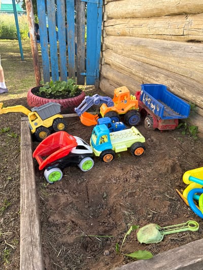 This is a medium-shot, eye-level, outdoor photograph taken in a sandbox at a property in Chuy-Atasevo, Russia. The sandbox is bordered by wooden planks and contains several toy construction vehicles, including a yellow and grey dump truck with an excavator arm, a red and black dump truck, a light green and blue dump truck, an orange and blue digger, and a blue dump truck. Scattered around the sandbox are a green plastic shovel and a yellow and blue watering can. In the background, there is a wooden fence and a small garden bed planted with seedlings in a red tire. The ground is covered in dirt and dry grass.